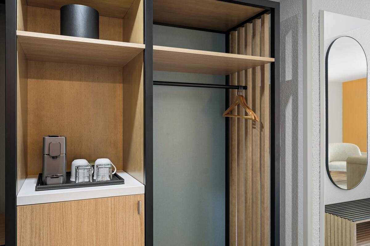 A modern hotel room with a wooden cabinet, a coffee maker, and a hanging coat rack.