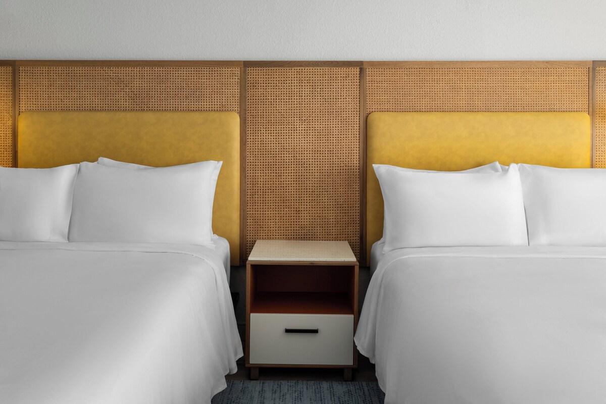 A hotel room with two beds, a wooden nightstand with a drawer, and a woven headboard.