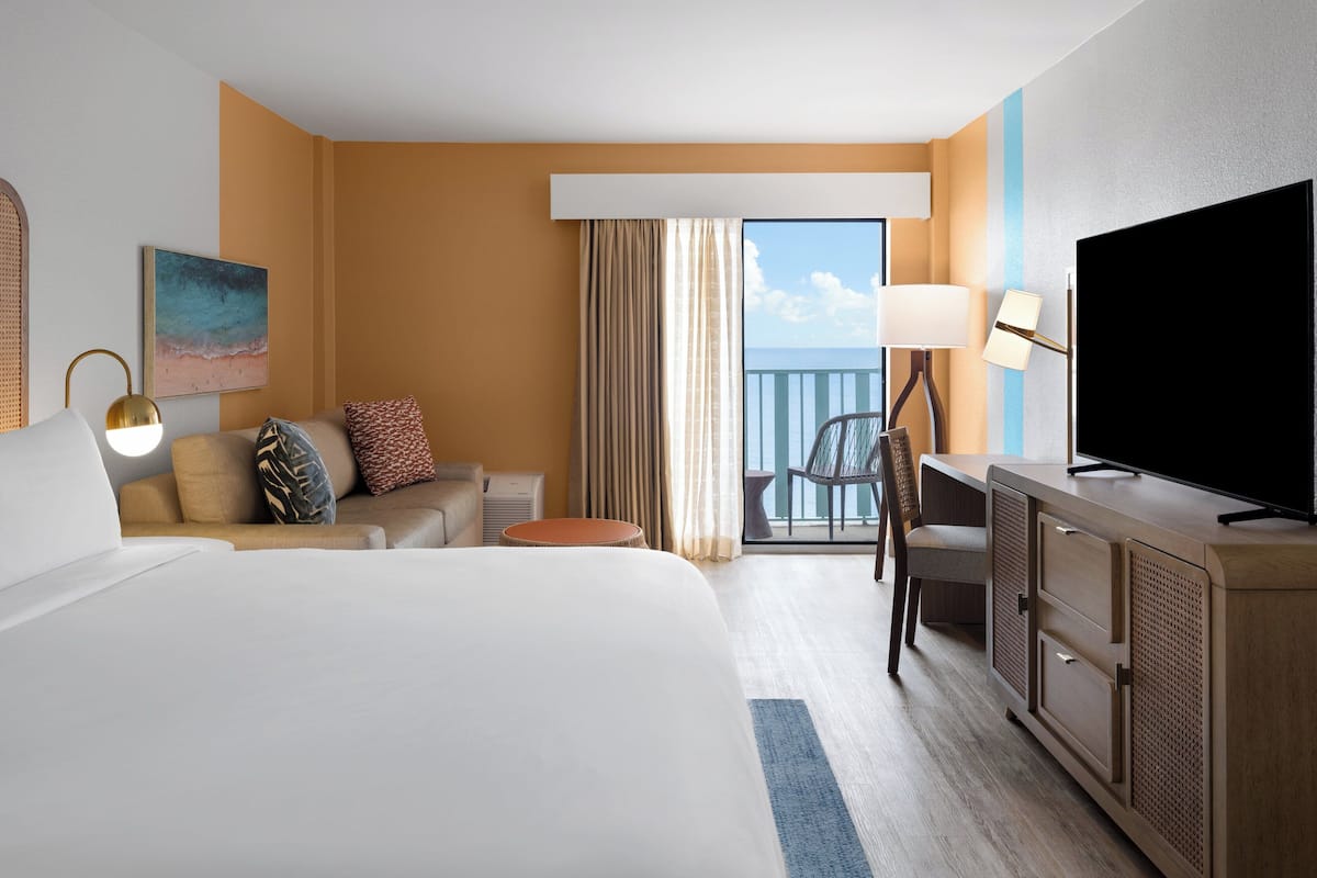 A hotel room with a large bed, a desk with a TV, a chair, a sofa with pillows, a small table, and a window with curtains.