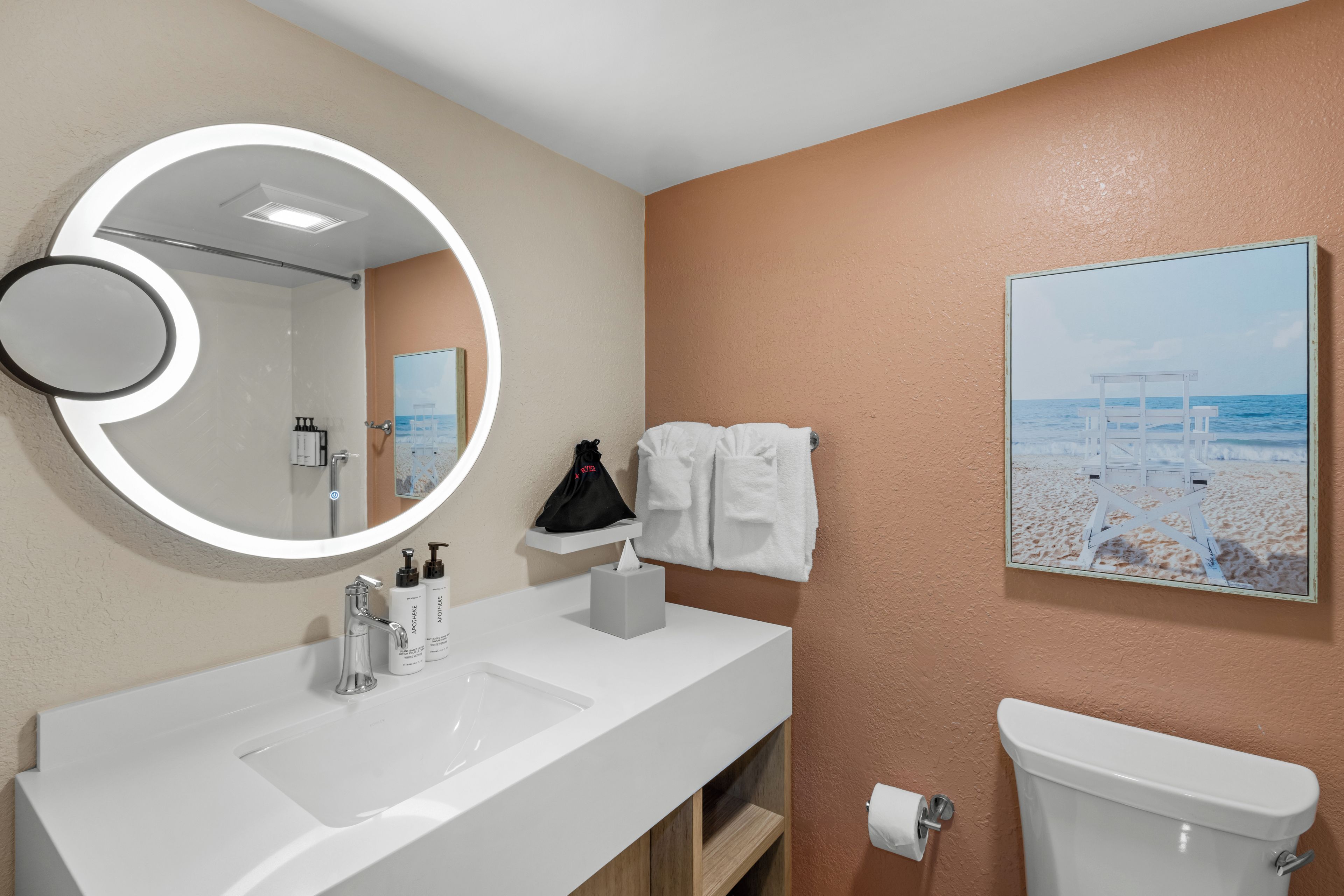 A bathroom with a white sink, a toilet, a towel rack, a mirror with a circular light, and a beach-themed painting on the wall.