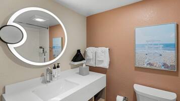 A bathroom with a white sink, a toilet, a towel rack, a mirror with a circular light, and a beach-themed painting on the wall.