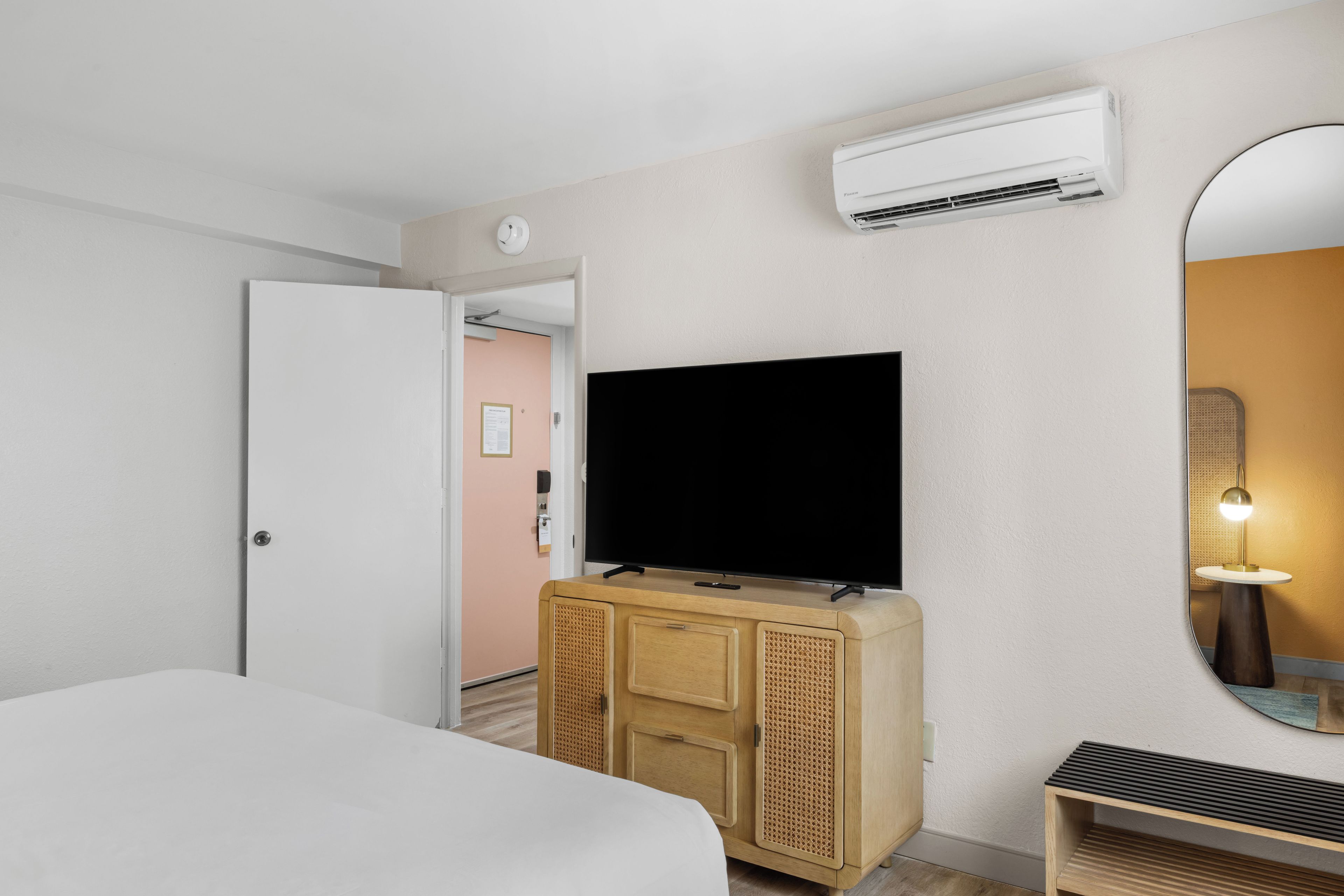Une chambre d’hôtel avec un lit, un téléviseur posé sur une commode en bois, un appareil de climatisation, une porte et un miroir.