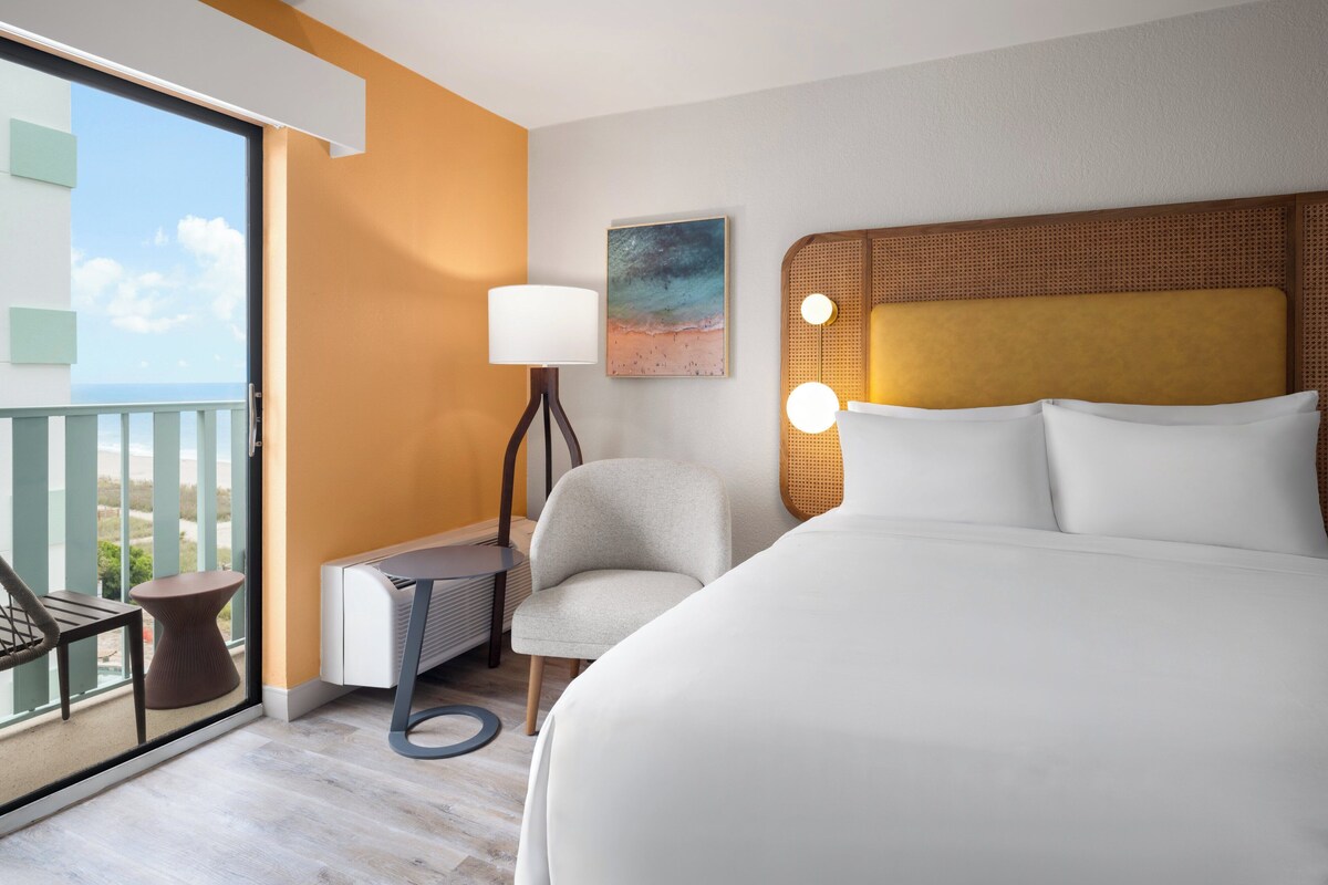 A hotel room with a large bed, a chair, a small table, a lamp, and a painting on the wall.