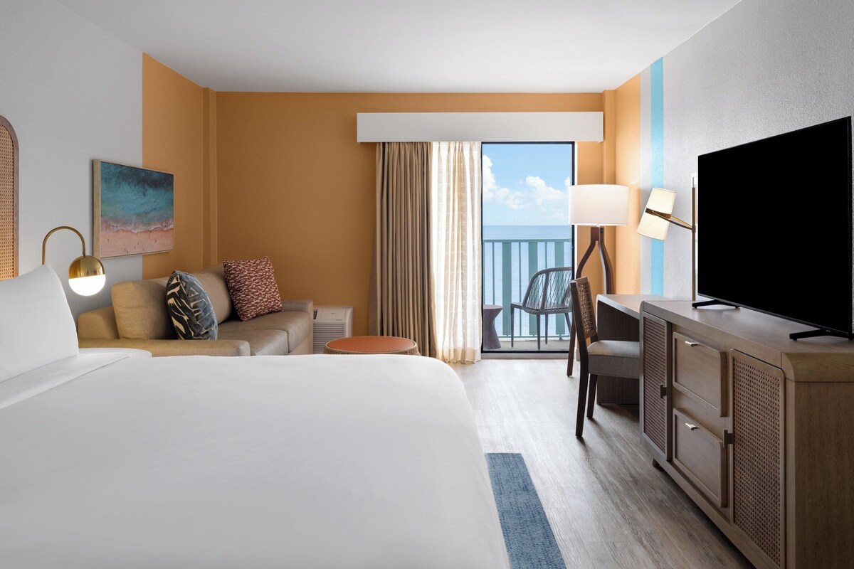 A hotel room with a large bed, a desk with a TV, a chair, a sofa with pillows, a small table, and a window with curtains.