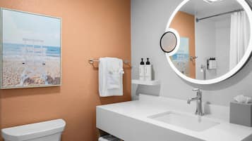 A bathroom with a white sink, a round mirror, a towel rack with a white towel, and a framed picture of a beach scene.