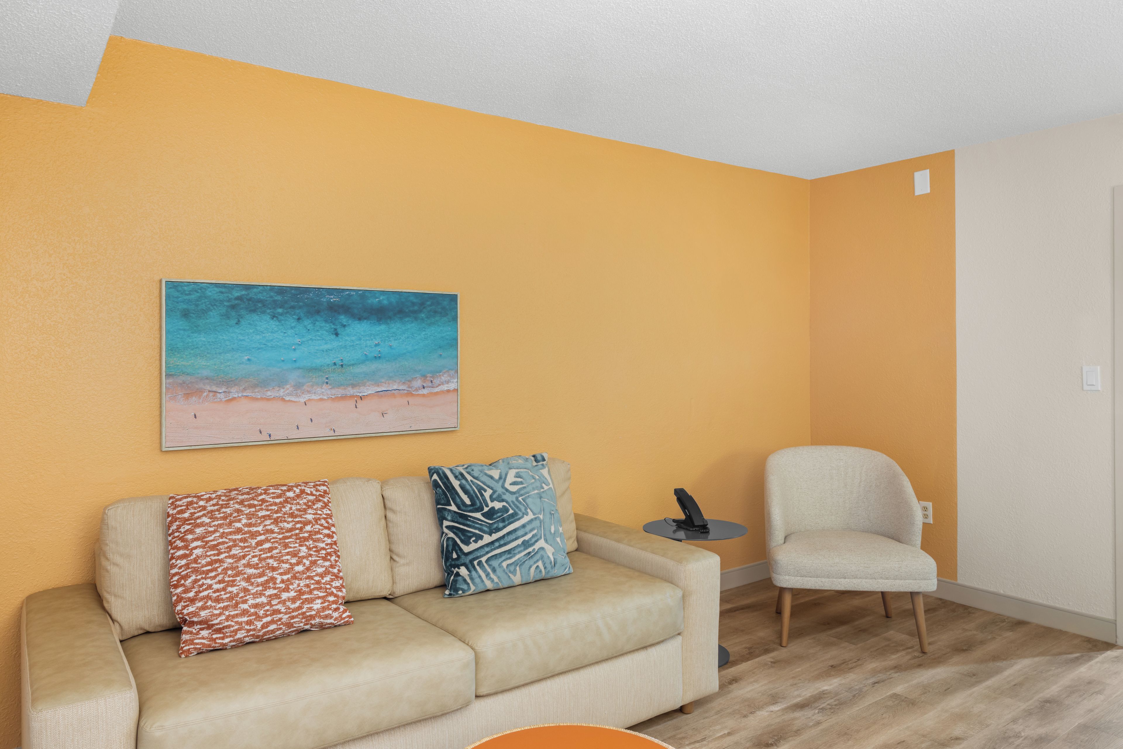 A living room with a beige sofa, a blue and white patterned pillow, a small round table with a black object, and a painting of a beach scene on the wall.
