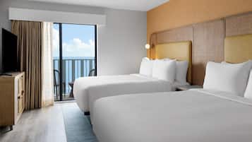 A hotel room with two beds, a TV, a desk, and a balcony with a view of the sea.