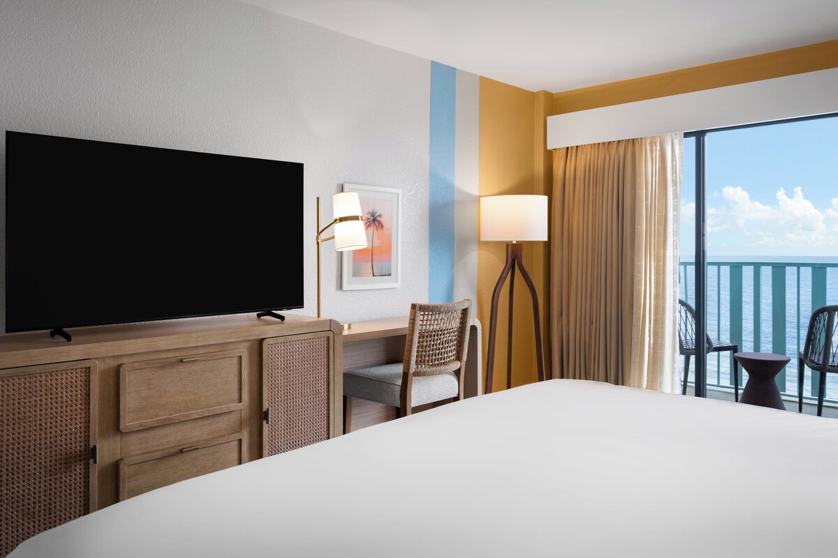 A hotel room with a large bed, a desk with a television, a chair, a lamp, and a view of the ocean through a balcony.