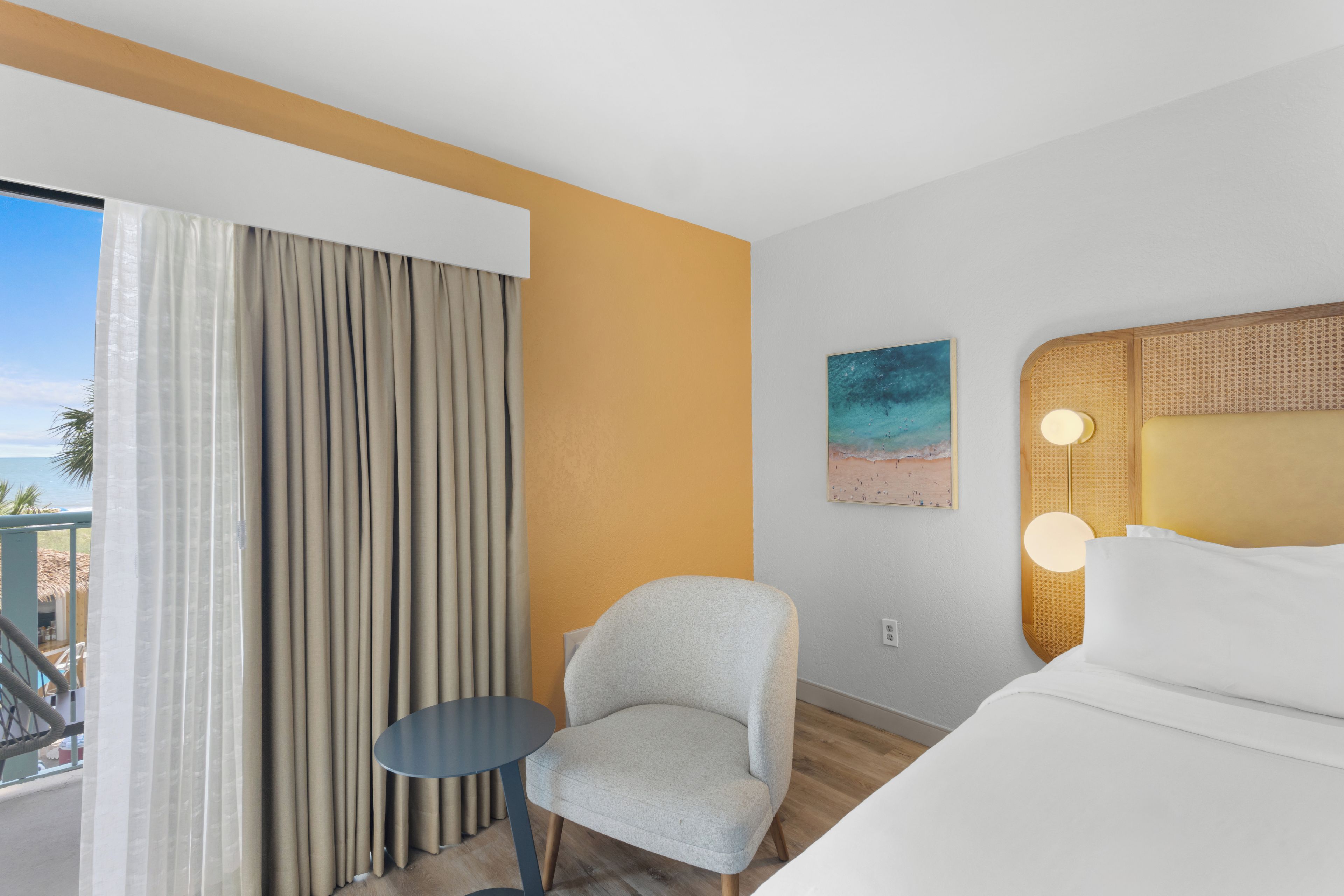 A hotel room with a bed, a chair, a small table, a painting, and a view of the outdoors.