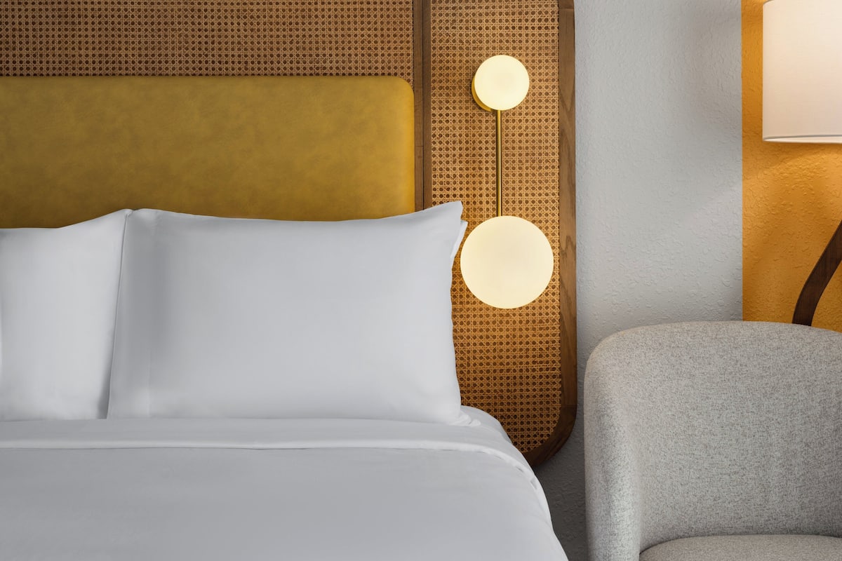 A neatly made bed with white linens, a round wall lamp, and a grey armchair.