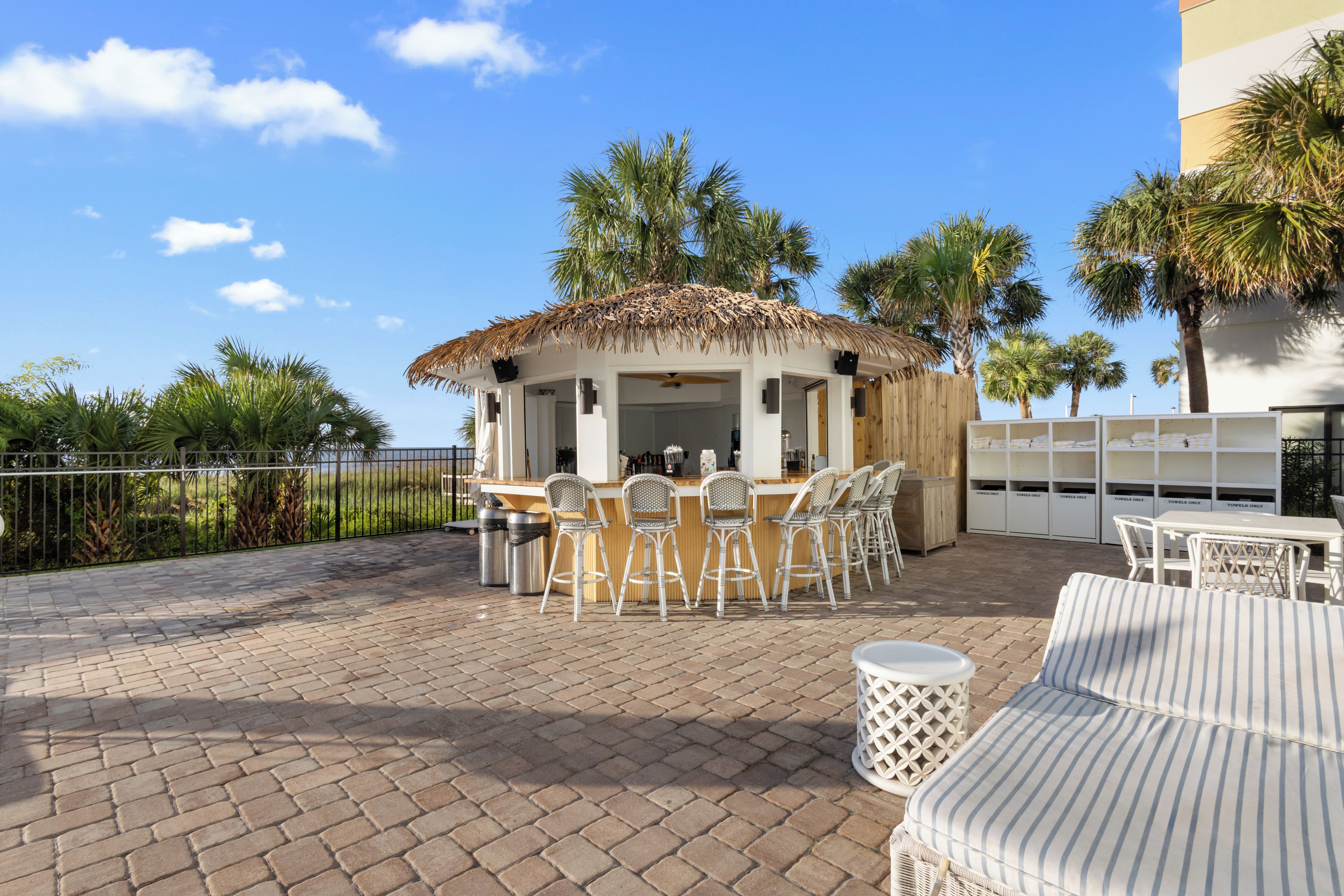 An outdoor bar area with a thatched roof, white wicker furniture, and a view of palm trees.