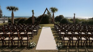 An outdoor wedding venue with a white aisle carpet, wooden chairs, and palm trees.