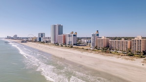 A coastal cityscape with a wide sandy beach, high-rise buildings, and a pier extending into the ocean.