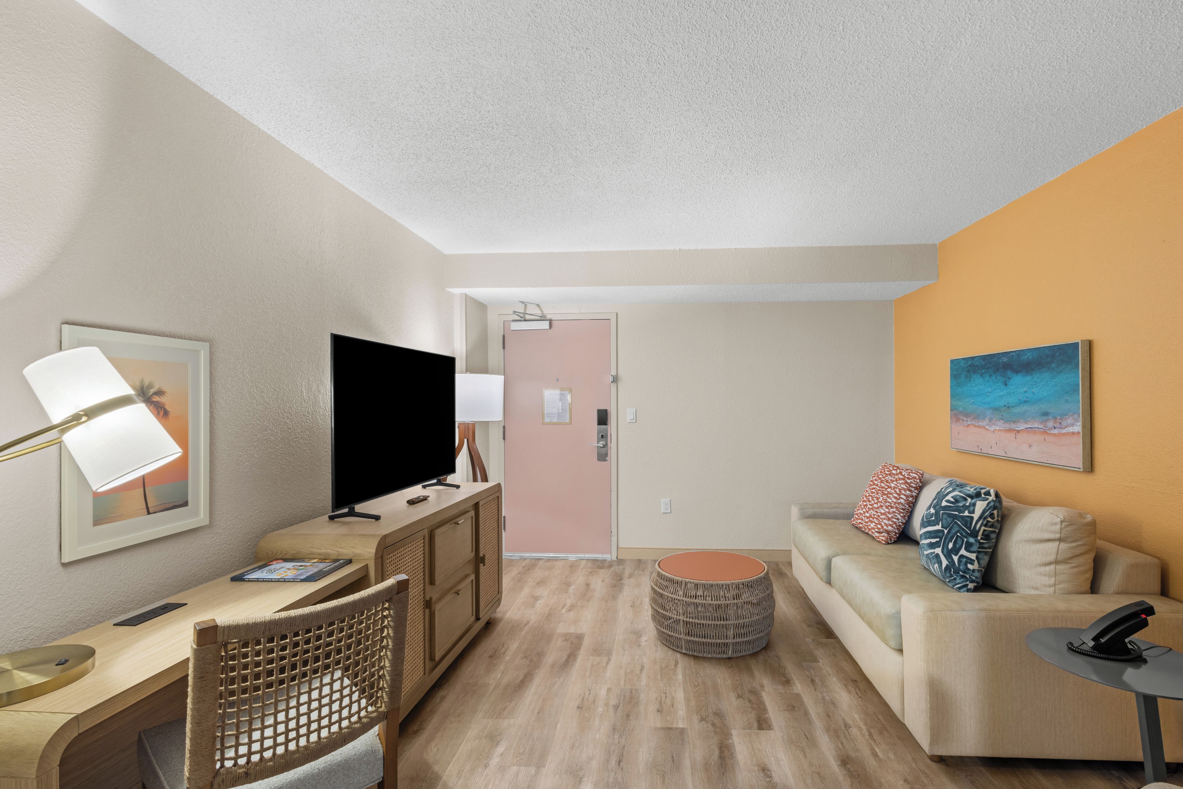 Un salon moderne avec un téléviseur à écran plat, un bureau avec une lampe, un canapé avec des coussins et un pouf rond.