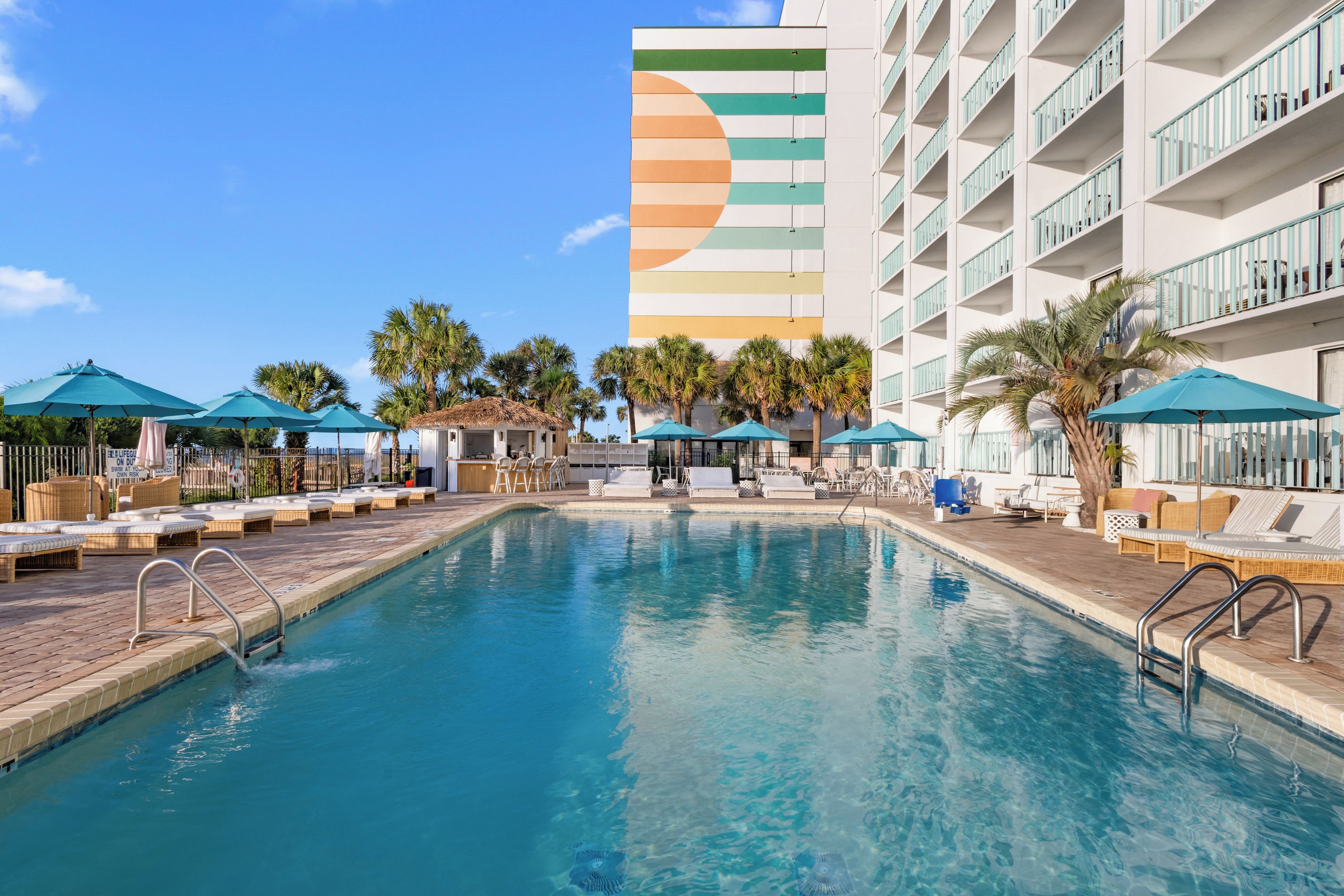 A swimming pool with lounge chairs and umbrellas, surrounded by palm trees and a multi-story building with a colorful facade.
