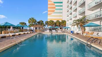 A swimming pool with lounge chairs and umbrellas, surrounded by palm trees and a multi-story building with a colorful facade.