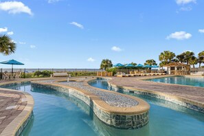 A swimming pool with a curved design, surrounded by lounge areas with umbrellas and palm trees.