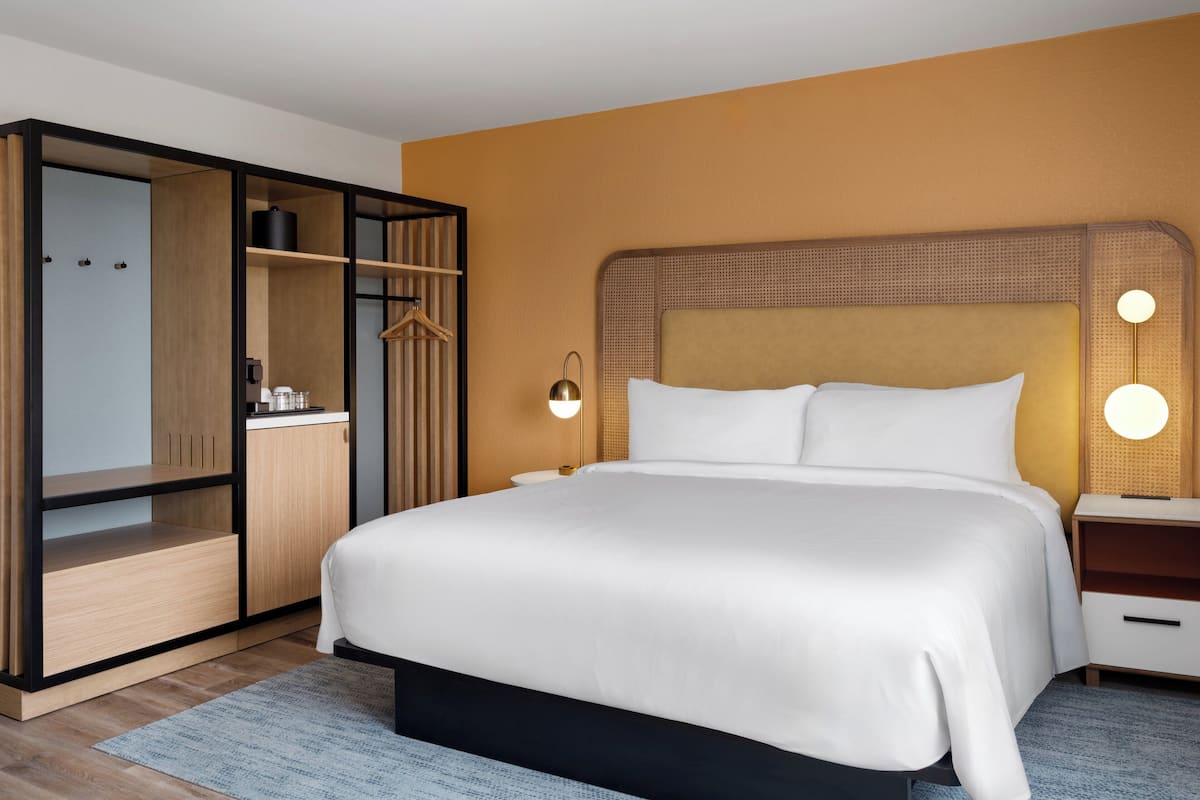 A modern hotel room with a large bed, built-in wardrobe, bedside tables, and wall-mounted lighting.