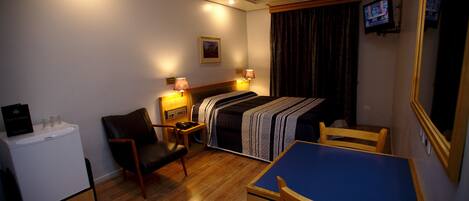 A compact hotel room with a bed, a desk, a chair, a small table, a refrigerator, and a television.