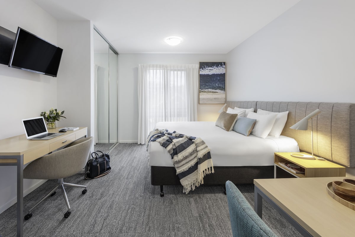 A modern hotel room with a bed, desk, chair, and a TV mounted on the wall.