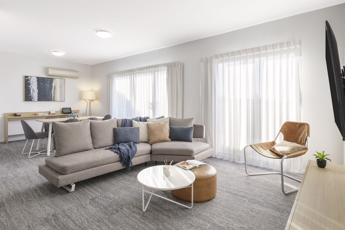 A modern living room with a sofa, coffee table, and armchair, a dining area with a table and chairs, and large windows with sheer curtains.