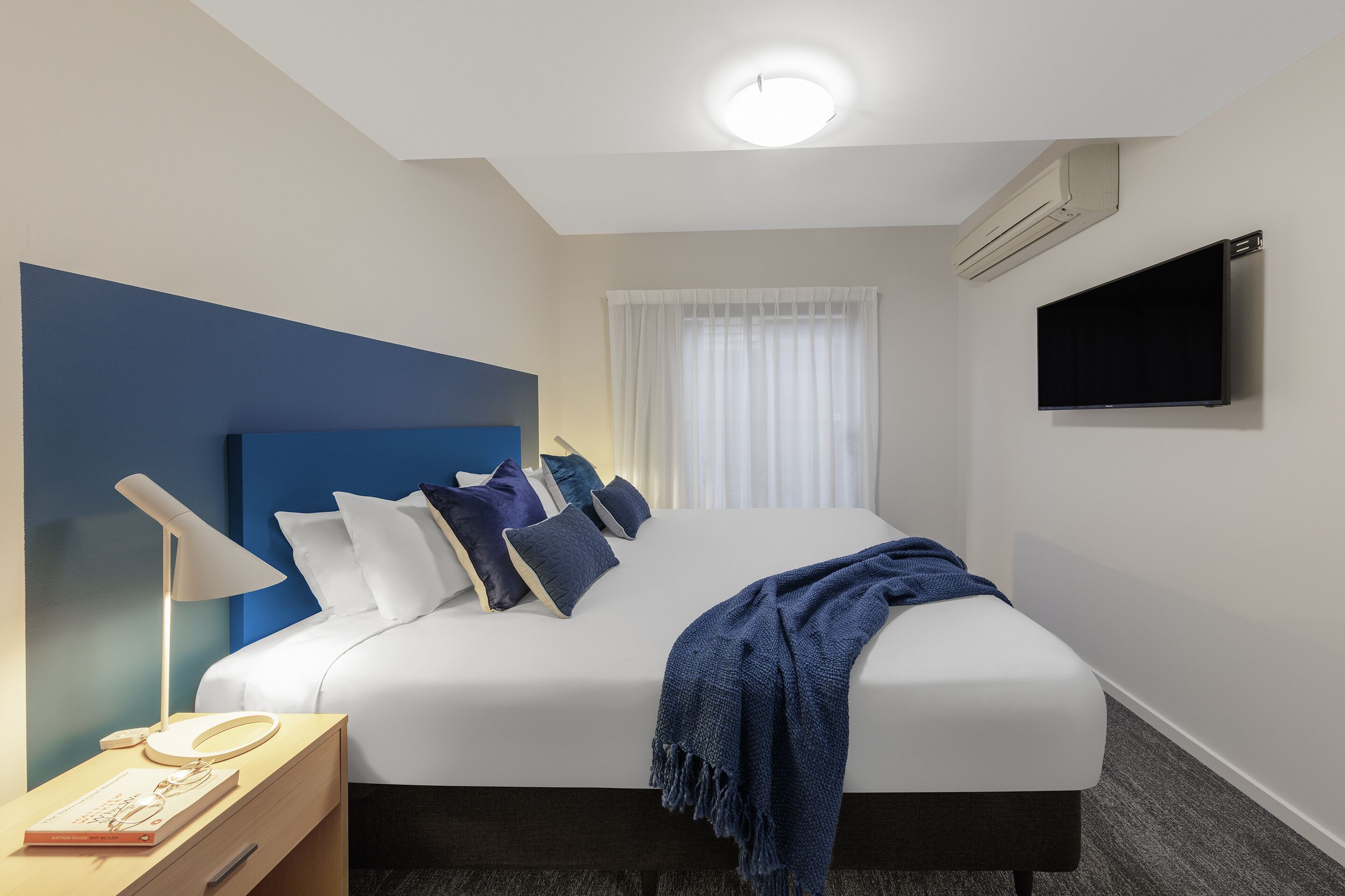 A hotel room with a bed, a bedside table with a lamp, a flat-screen TV mounted on the wall, and a window with sheer curtains.