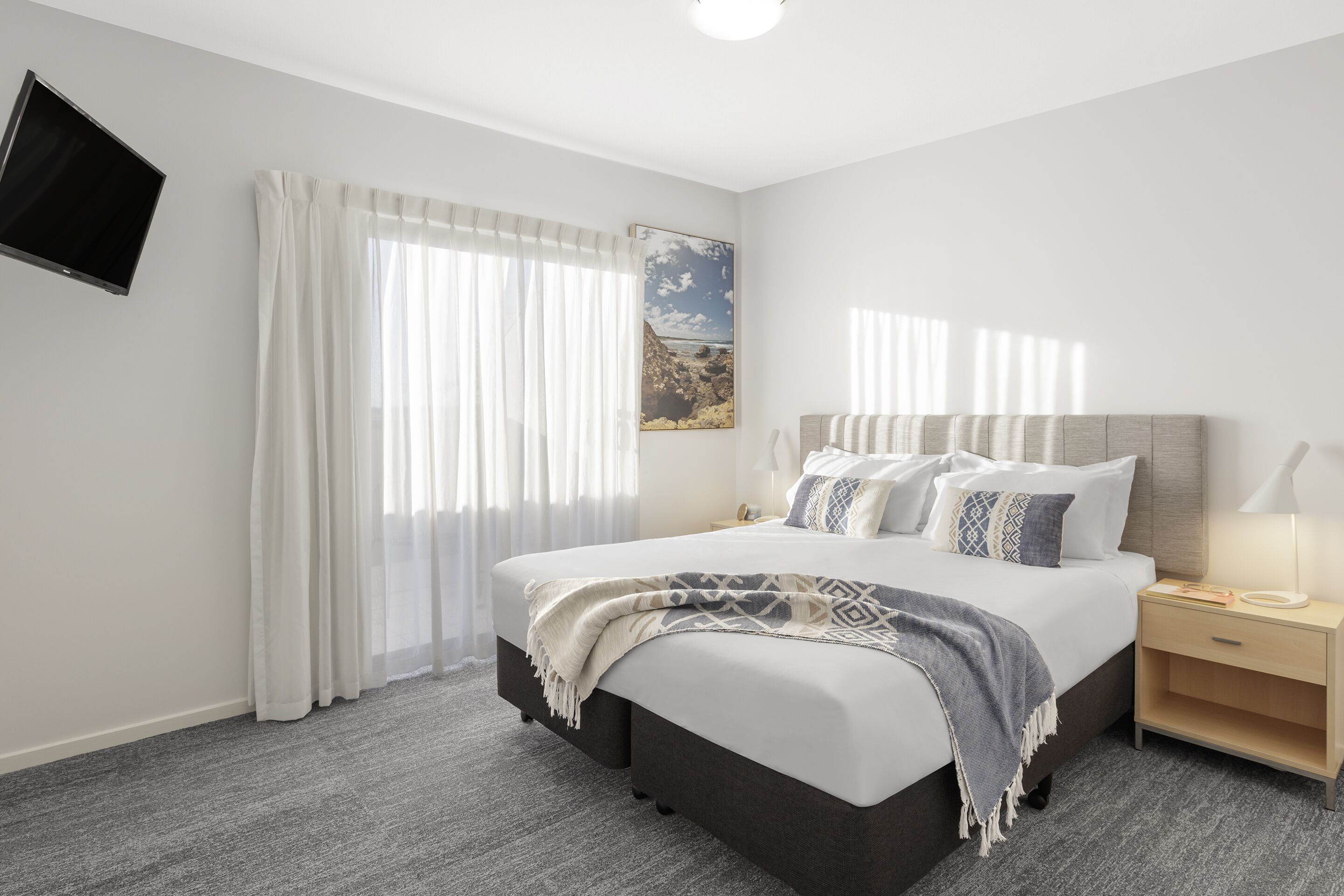 Une chambre d’hôtel moderne avec un grand lit, une table de chevet avec une lampe, un téléviseur à écran plat fixé au mur et une fenêtre avec des rideaux légers.