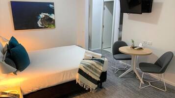 Quarto de hotel com cama, escrivaninha com luminária, cadeira, mesinha e televisão na parede.