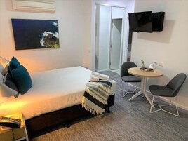 Habitación de hotel con cama, escritorio con lámpara, silla, una mesita y televisor montado en la pared.
