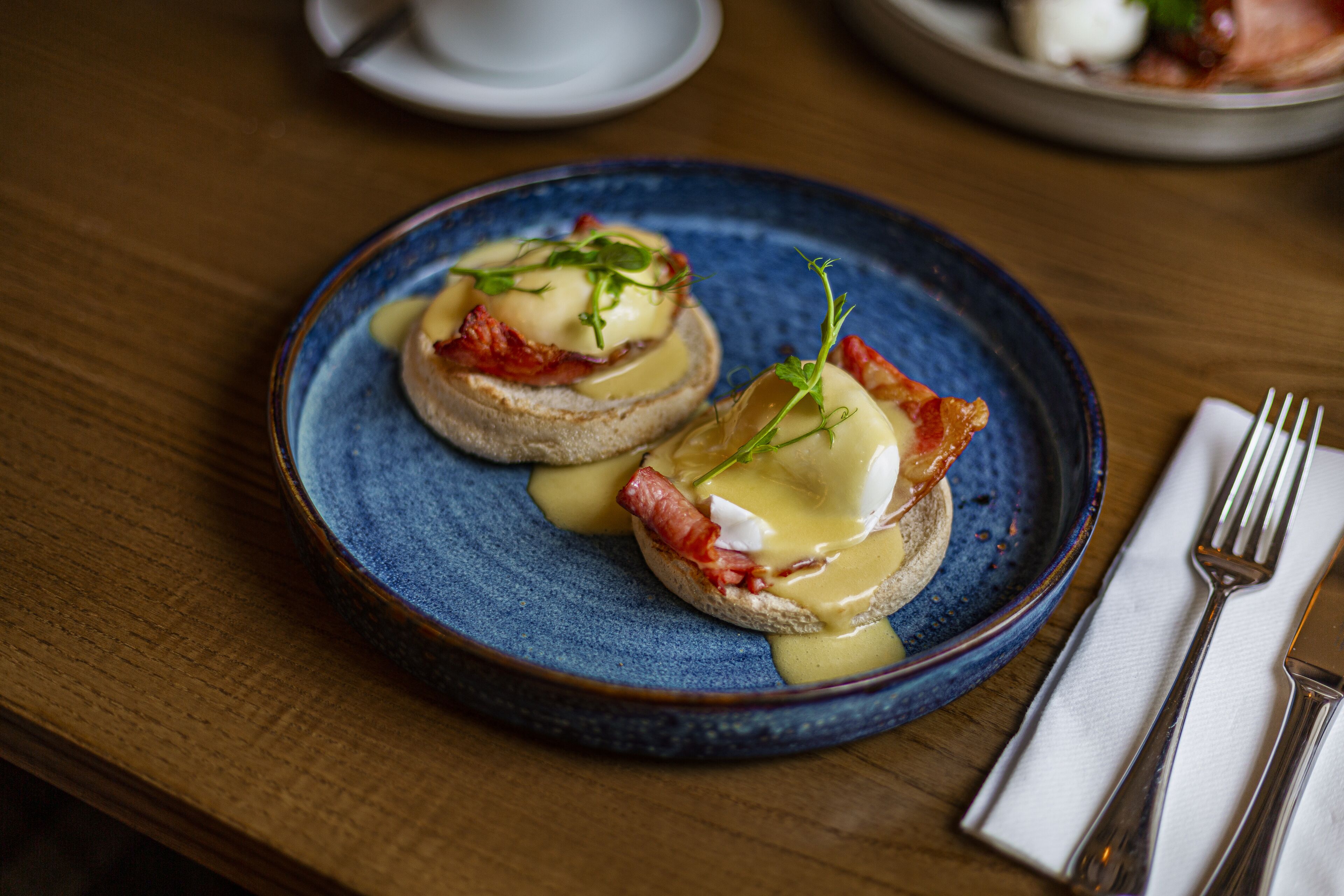 Two eggs benedict sandwiches on a blue plate with a fork and napkin on a wooden table.