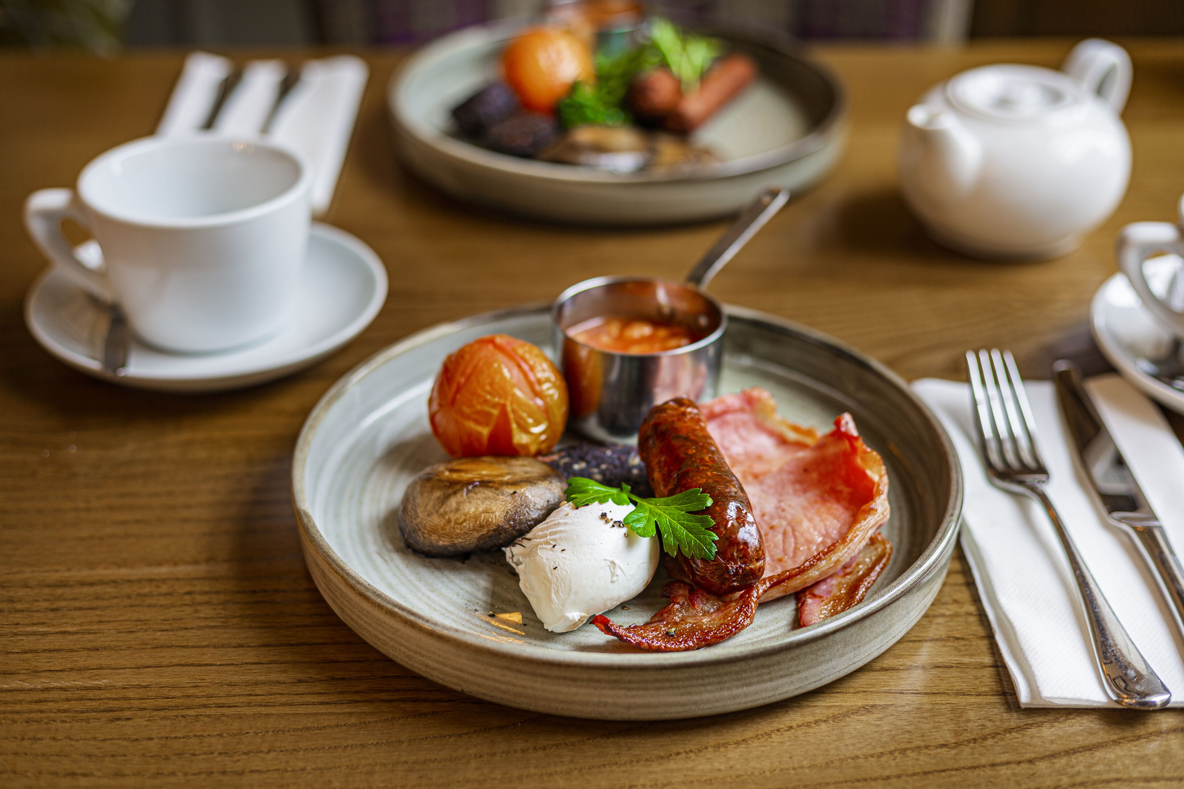 A breakfast setting with a plate of food, a cup, and a teapot on a wooden table.