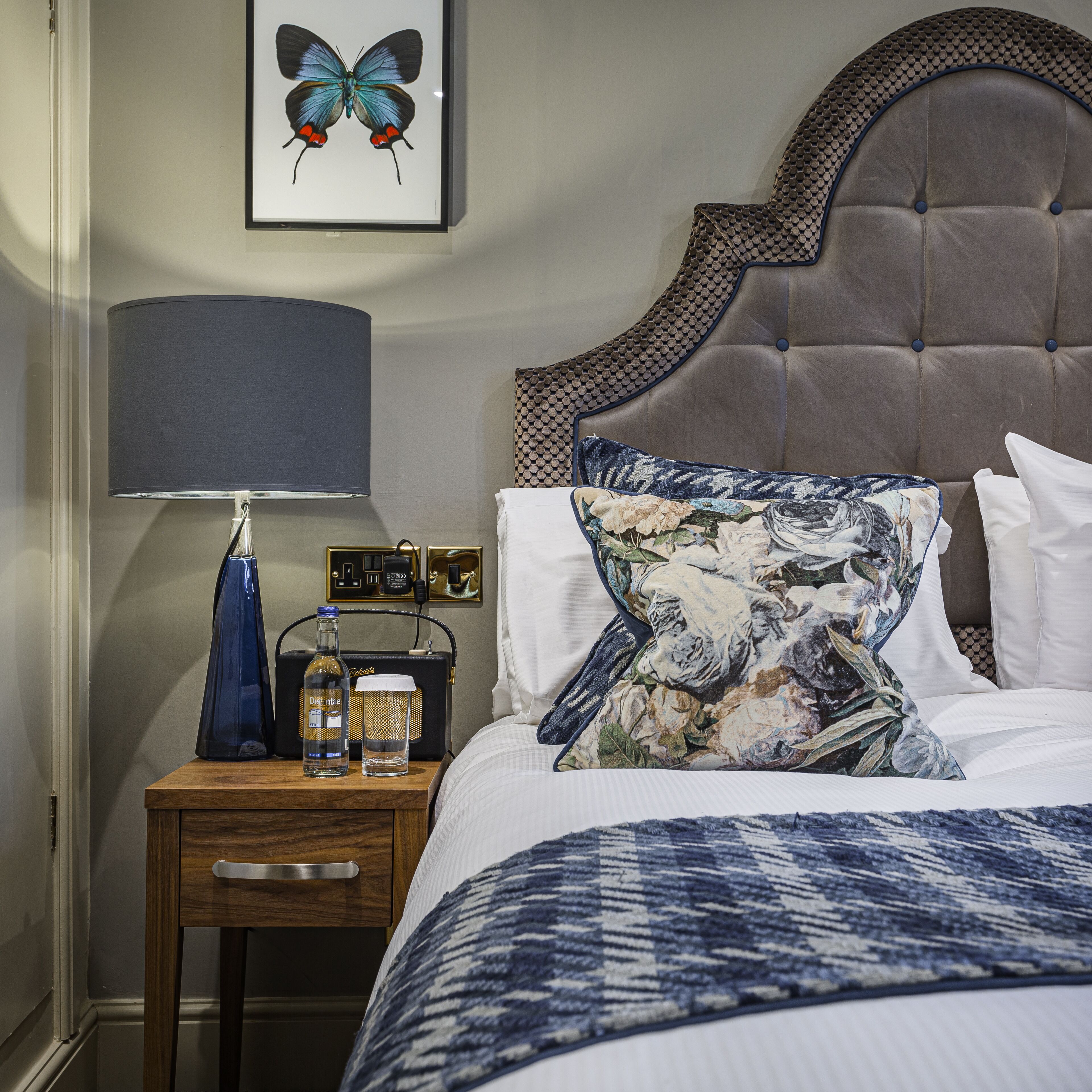 A neatly made bed with a patterned blanket, a decorative pillow, a bedside lamp, and a small table with a bottle and a radio.