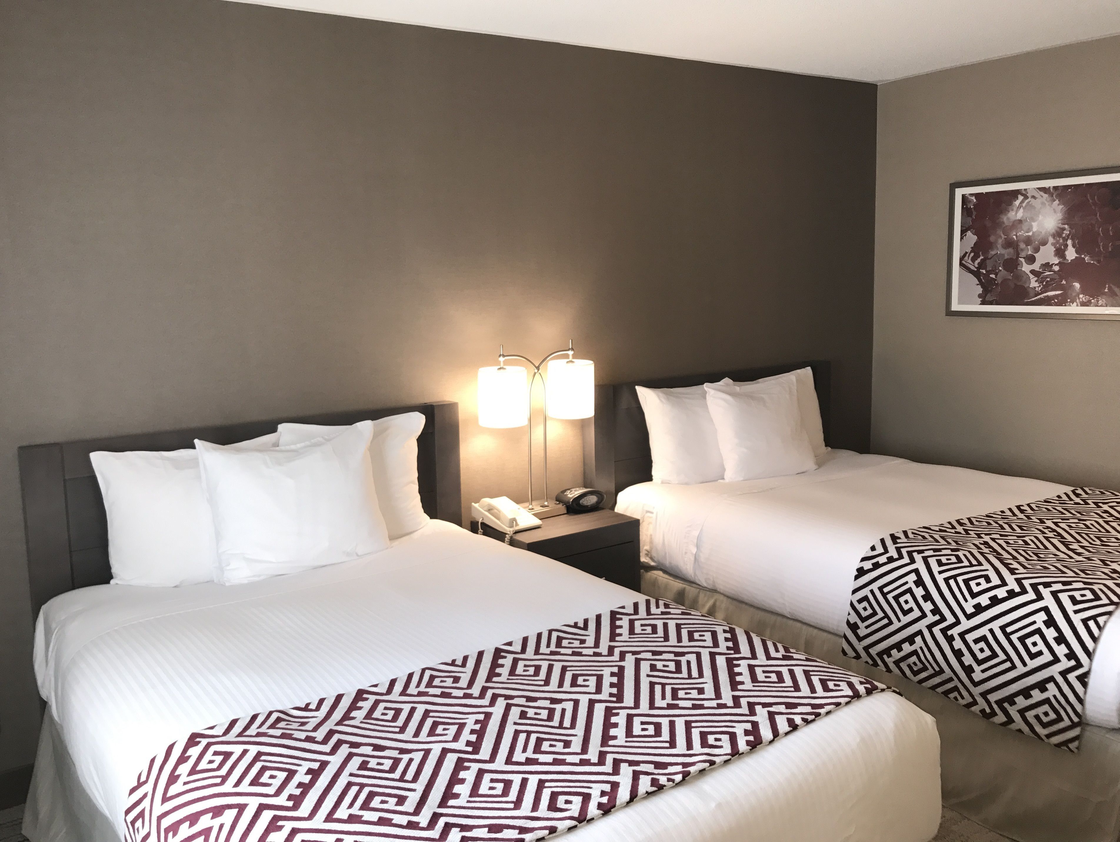 A hotel room with two beds, each with white bedding and patterned throw pillows. There are bedside lamps and a phone on the nightstand.