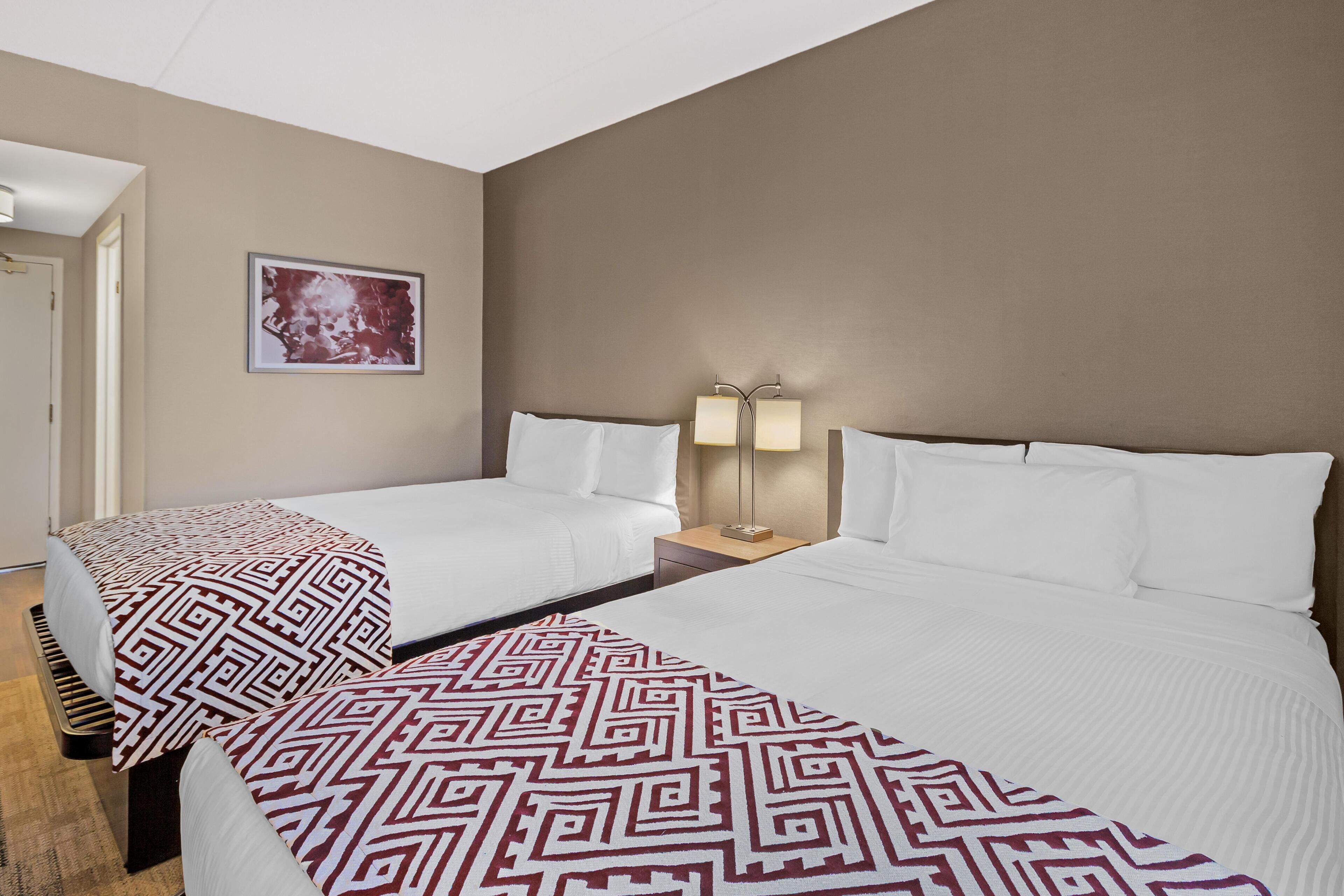 Habitación de hotel con dos camas, una mesita de noche con lámpara, un cuadro enmarcado en la pared y una puerta a la izquierda.