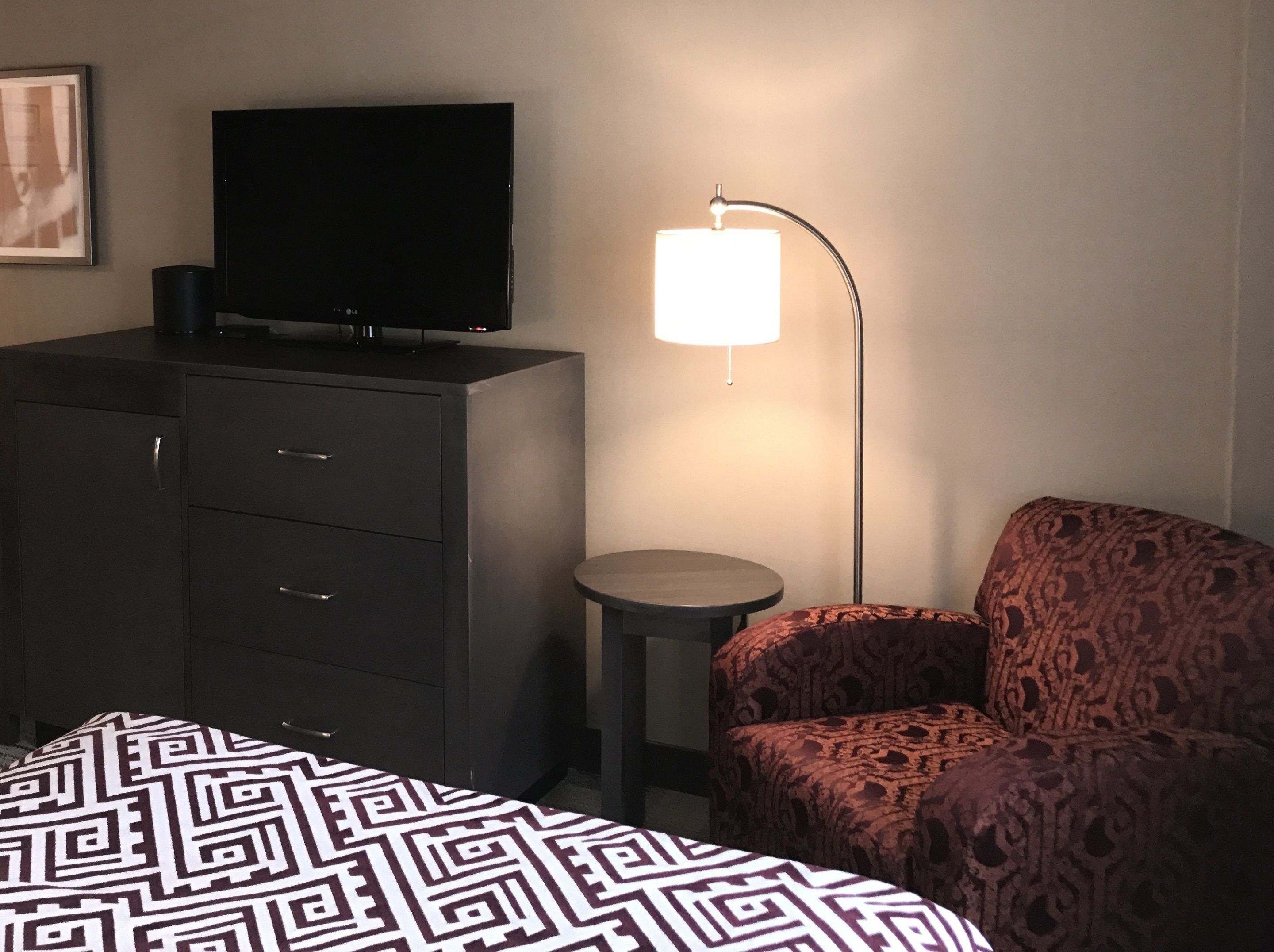 A hotel room with a bed, a TV on a dresser, a lamp, and a patterned armchair.