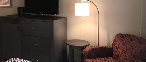 A hotel room with a bed, a TV on a dresser, a lamp, and a patterned armchair.