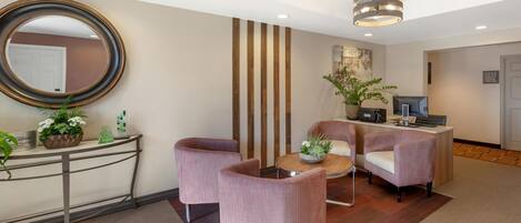 A reception area with a desk, two pink armchairs, a round coffee table, a large mirror, and a hanging light fixture.