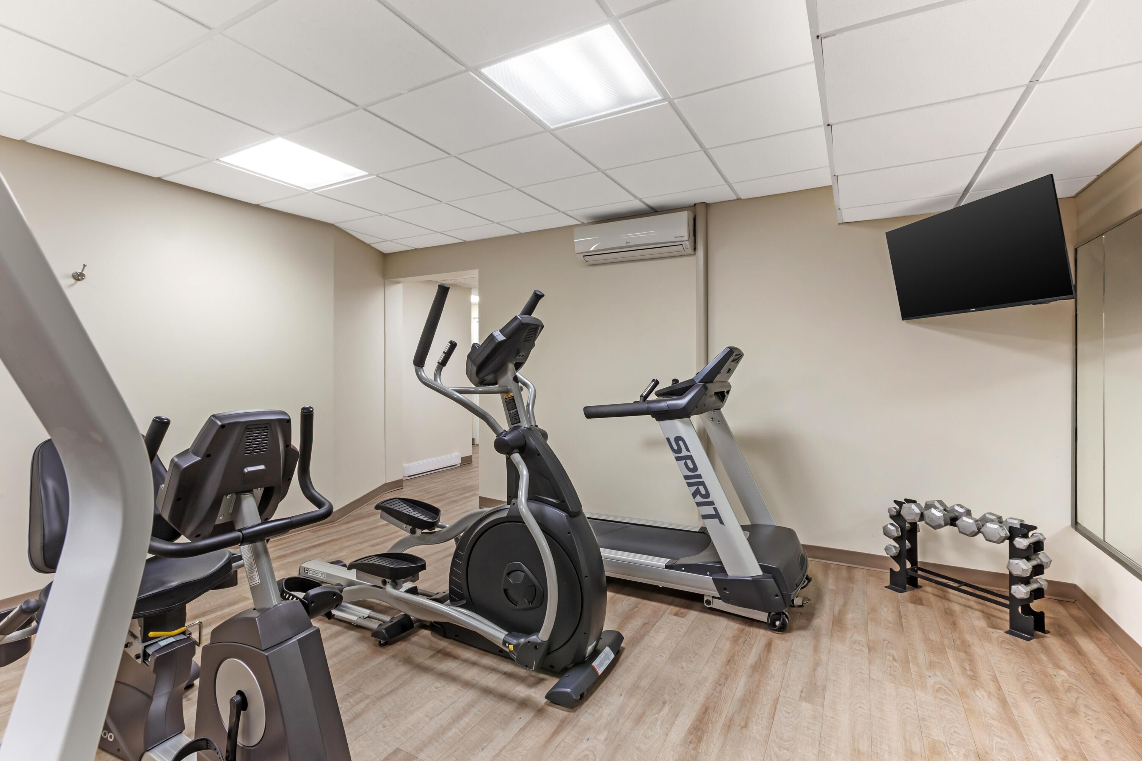 A gym with cardio machines, a wall-mounted TV, and a dumbbell rack.