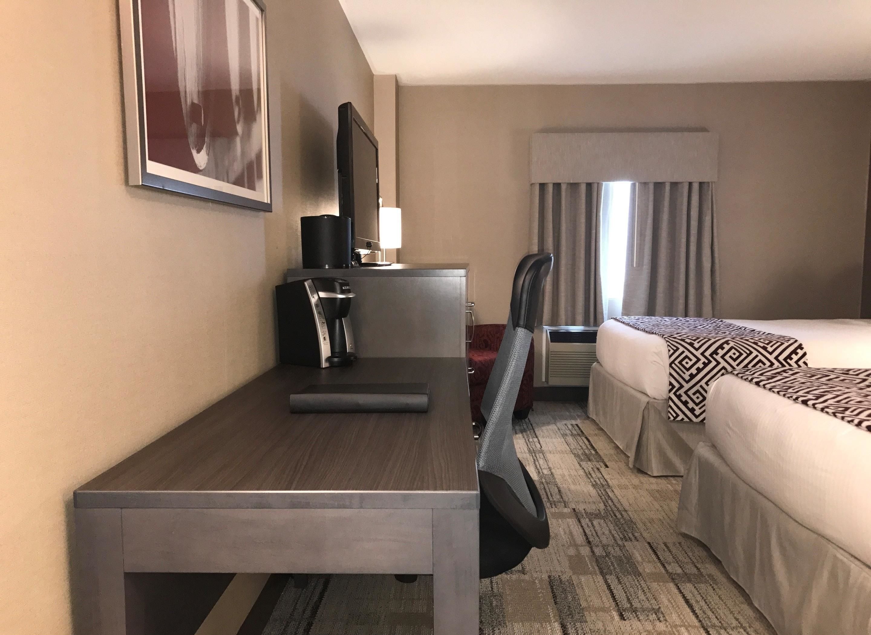 Una habitación de hotel con cama, escritorio con cafetera, silla, televisión y ventana con cortinas.