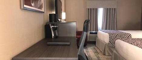 Una habitación de hotel con cama, escritorio con cafetera, silla, televisión y ventana con cortinas.