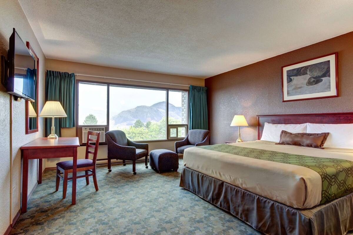 A hotel room with a bed, two chairs, a desk, a TV, and a large window with a mountain view.