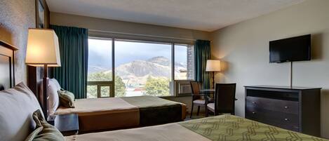 A hotel room with two beds, a desk, a chair, a TV, and a large window offering a mountain view.