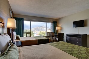 A hotel room with two beds, a desk, a chair, a TV, and a large window offering a mountain view.