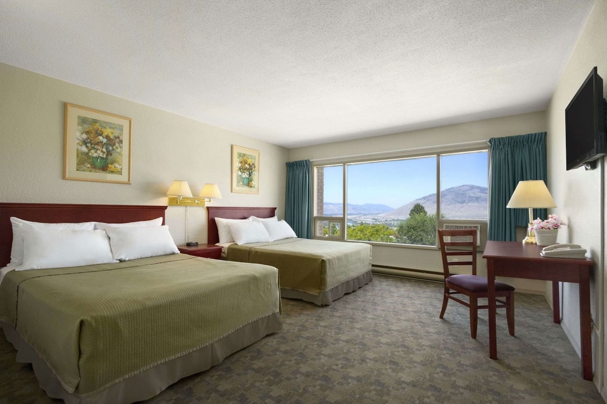 A hotel room with two beds, a desk, a chair, a TV, and a large window with a mountain view.