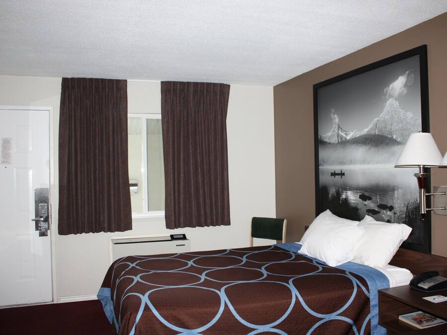 Een hotelkamer met een bed, nachtkastje, lamp, schilderij aan de muur en een raam met gordijnen.