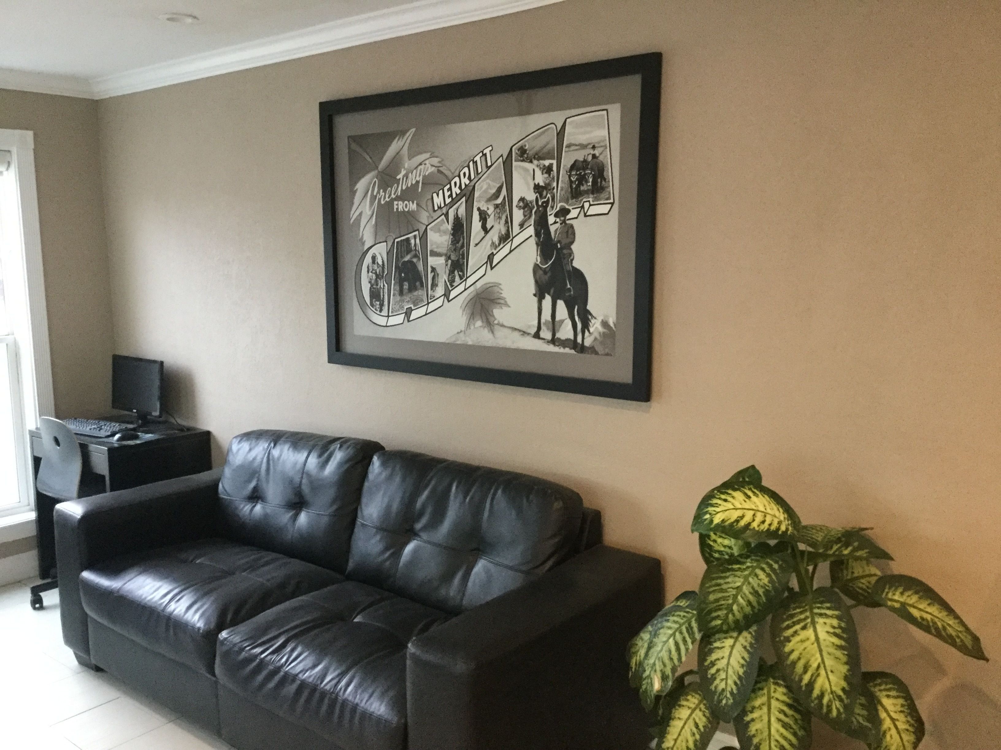 Un salon avec un canapé en cuir noir, un tableau encadré au mur, un bureau avec un ordinateur et une plante en pot.