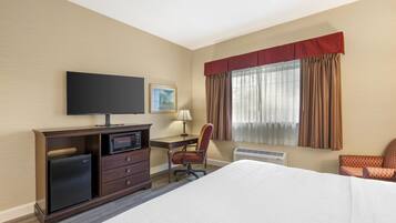 A hotel room with a bed, a desk with a computer, a television, a chair, a small refrigerator, and a window with curtains.