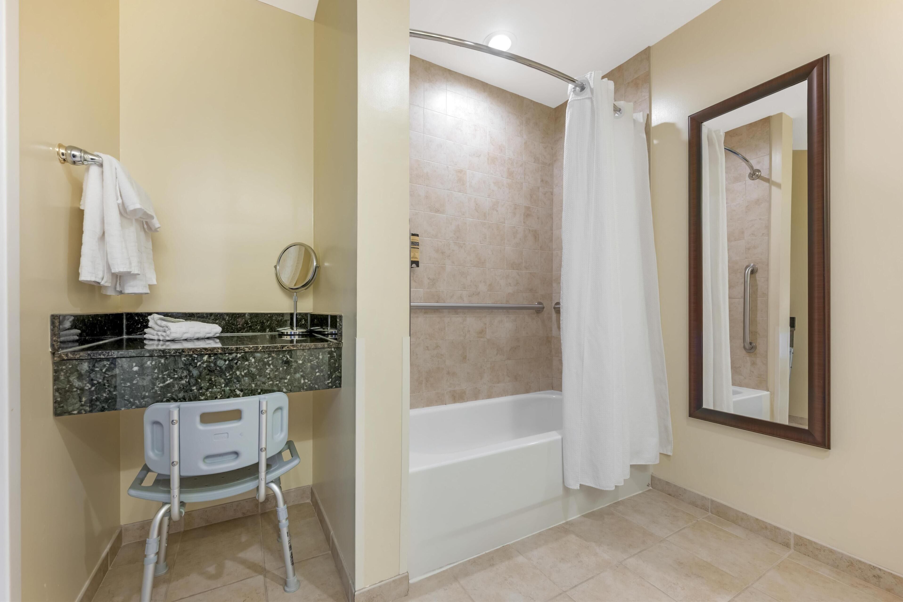 Un baño con ducha, inodoro con asiento accesible para silla de ruedas, lavamanos con encimera de mármol y espejo.