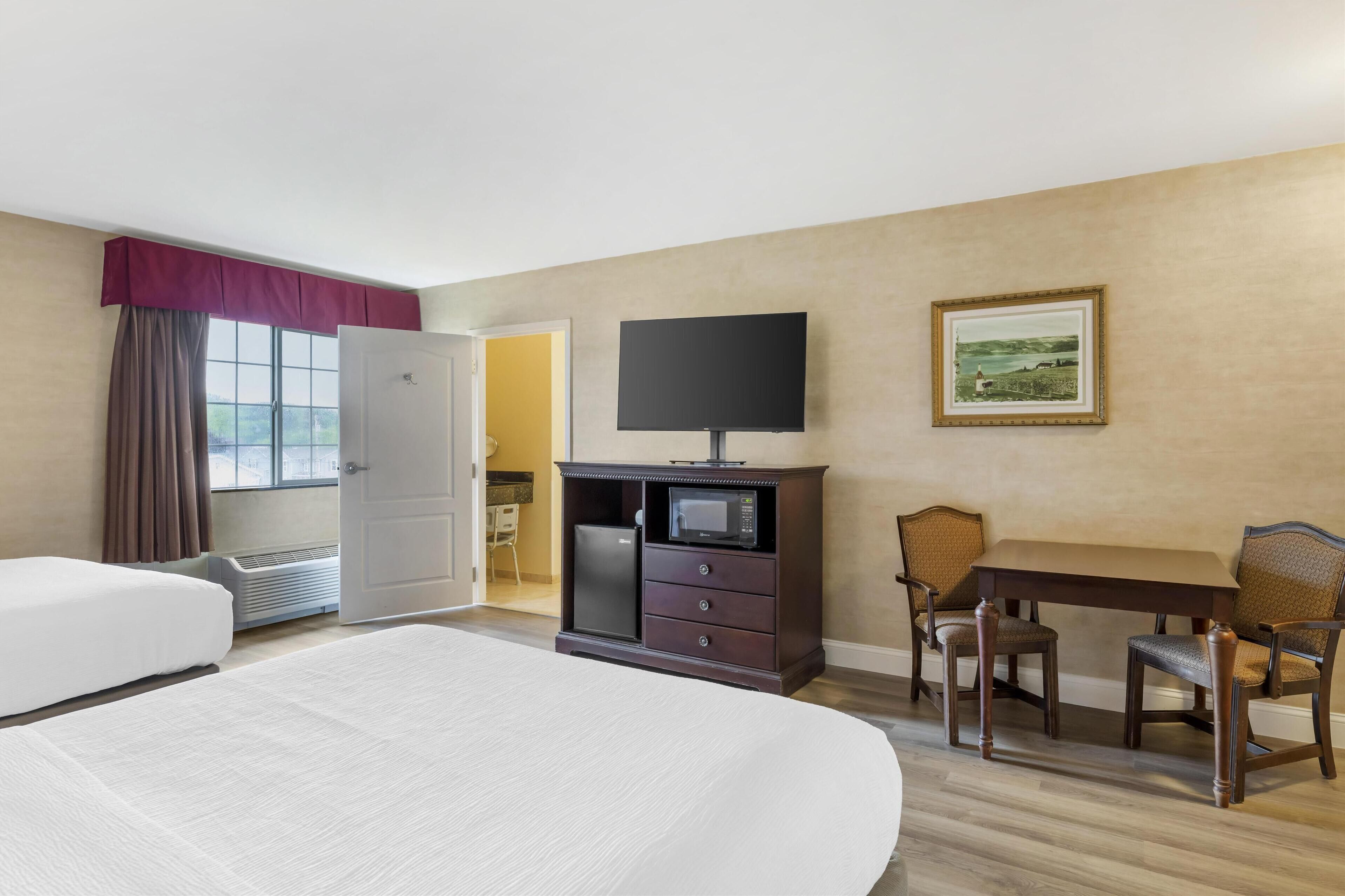 A hotel room with a bed, a TV, a microwave, a dining table with chairs, and a painting on the wall.
