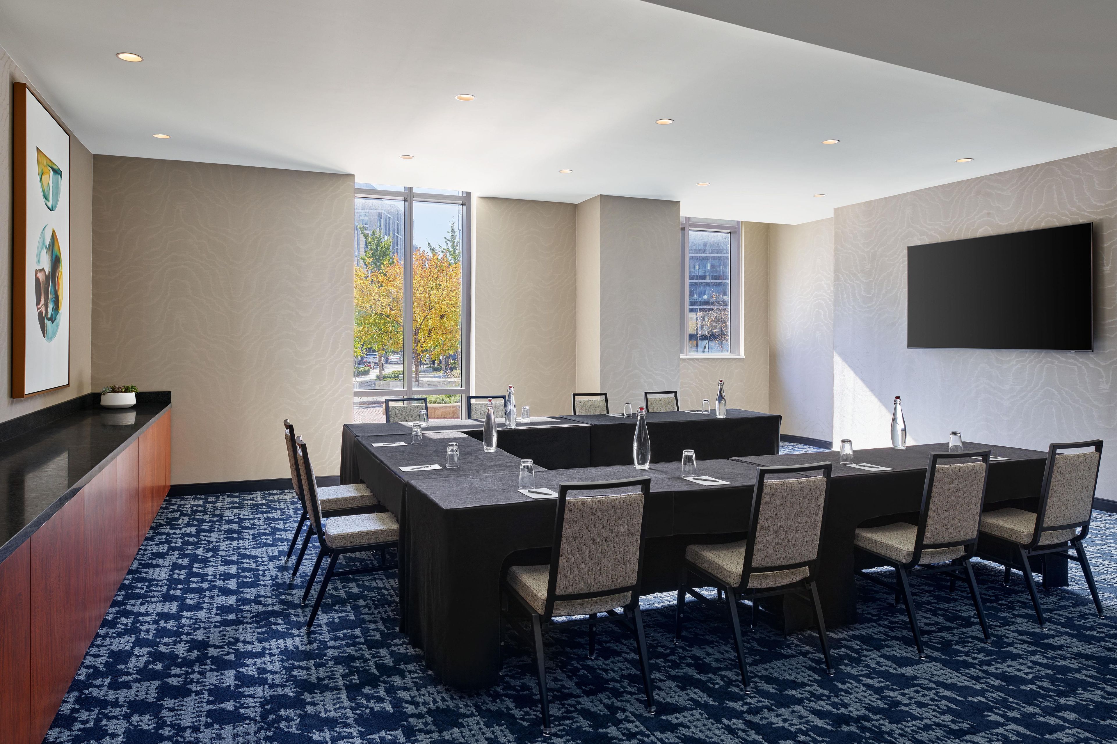 A conference room with a long table, chairs, a reception desk, and a large window with a view of trees and buildings.